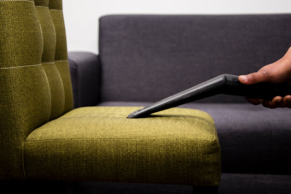 A person using a handheld vacuum cleaner to thoroughly clean a yellow and white patterned cushion placed on a dark fabric sofa in a residential living room, demonstrating surface cleaning and deep cle