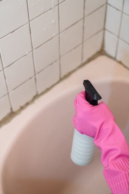 A close-up image showing a hand wearing a bright pink waterproof rubber glove, holding a white spray bottle with a black trigger, in the act of cleaning a white ceramic bathtub. The bathtub has a smoo